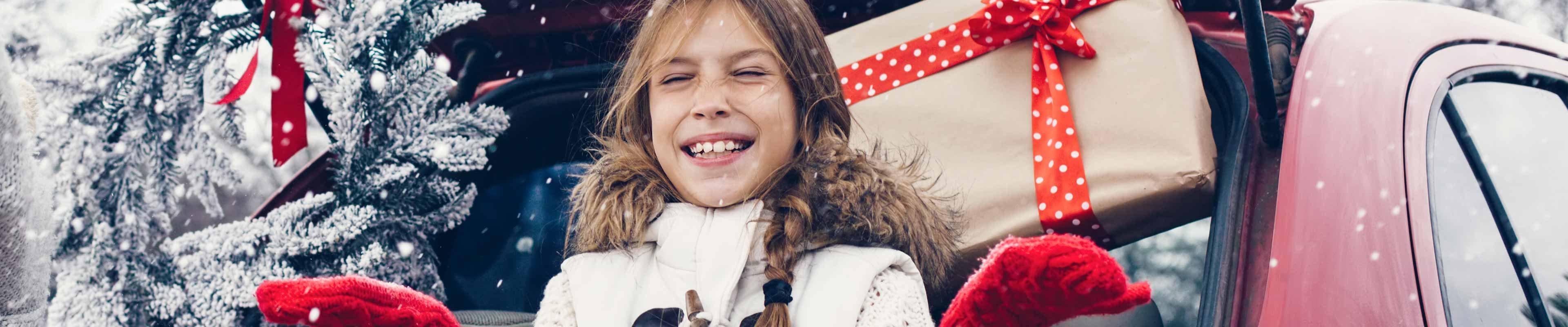 Two young, white children dressed for the holidays throwing snow in the air in front of a red car with its trunk open, revealing holiday presents.