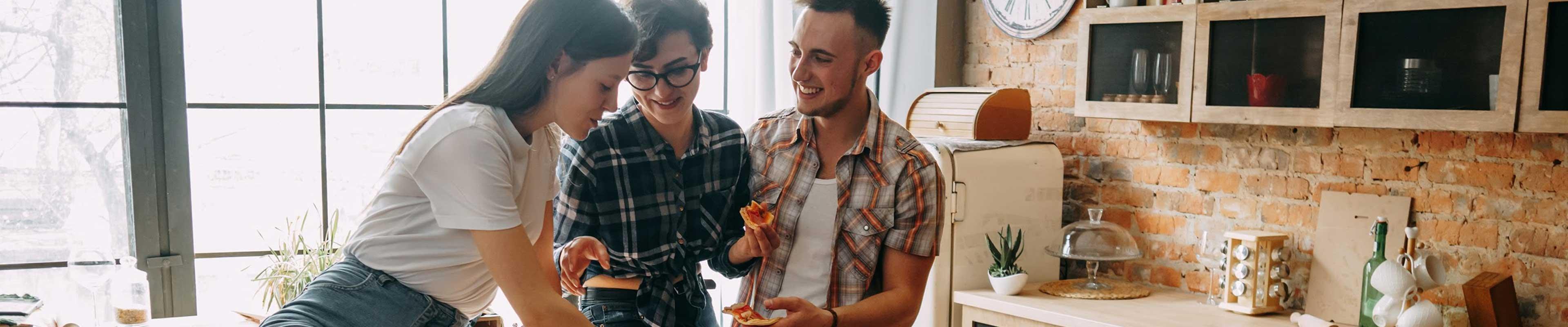 Three friends, two white women and one white man, sitting around a sunny kitchen eating homemade pizza and smiling.