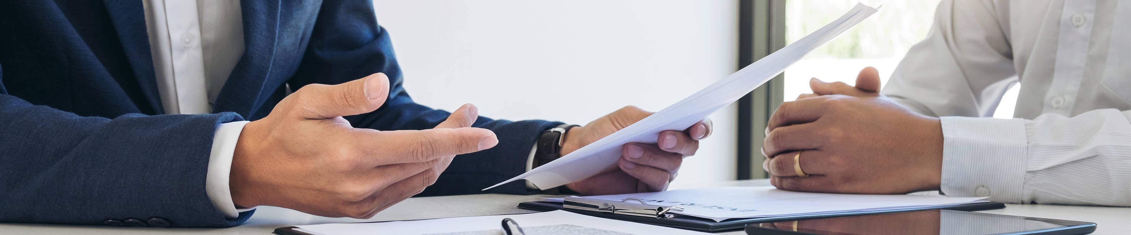 A white man holding a piece of paper explains insurance to a white woman with her hands folded on the desk.